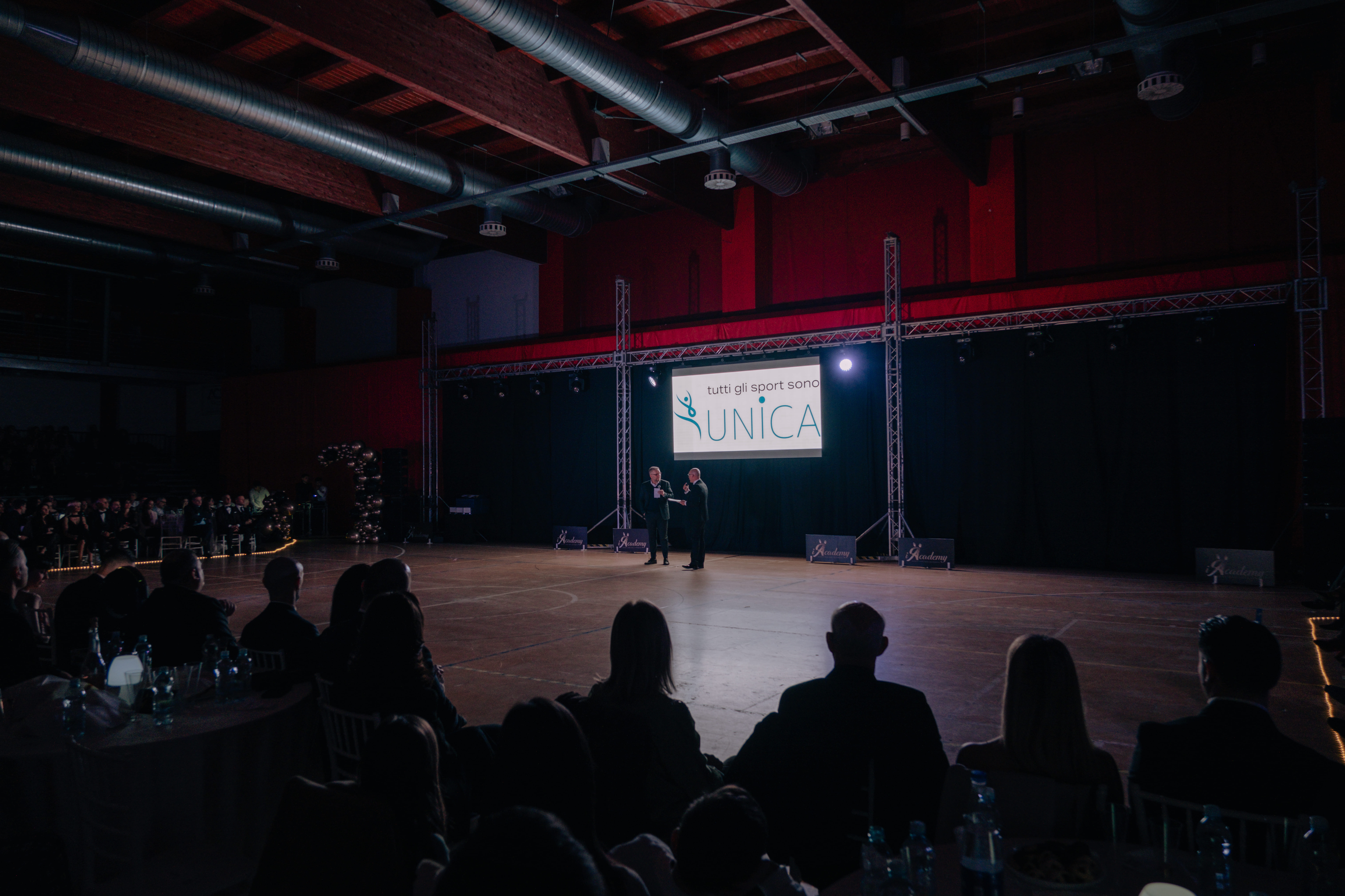 L'immagine ritrae due persone al centro del palco in un palazzetto: sono il Presidente di Unica e il presentatore della serata di gala. Di fronte a loro il palazzetto dello sport di Pomezia gremito.